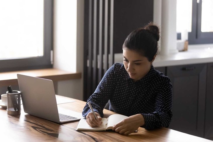 Concentrated millennial Indian woman sit at desk at home make notes study distant on laptop. Focused young ethnic female employee watch webinar take online course or work on internet on computer.